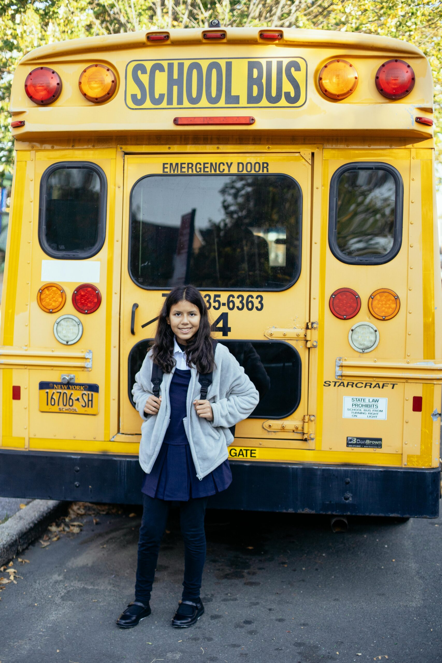 Content ethnic child in uniform near bright school bus looking at camera on asphalt roadway in town